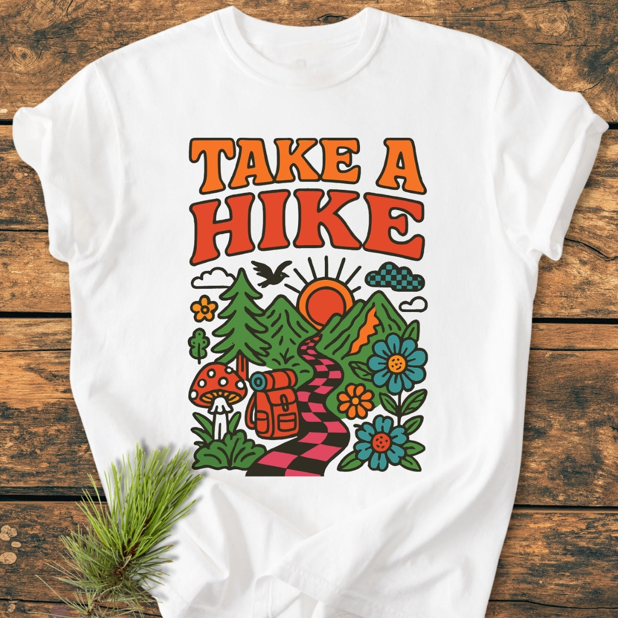 Take A Hike Funky - Vintage Wash Tee: A white T-shirt featuring a retro “TAKE A HIKE” design with a vibrant outdoor scene, laid on a wooden surface with a pine sprig beside it.