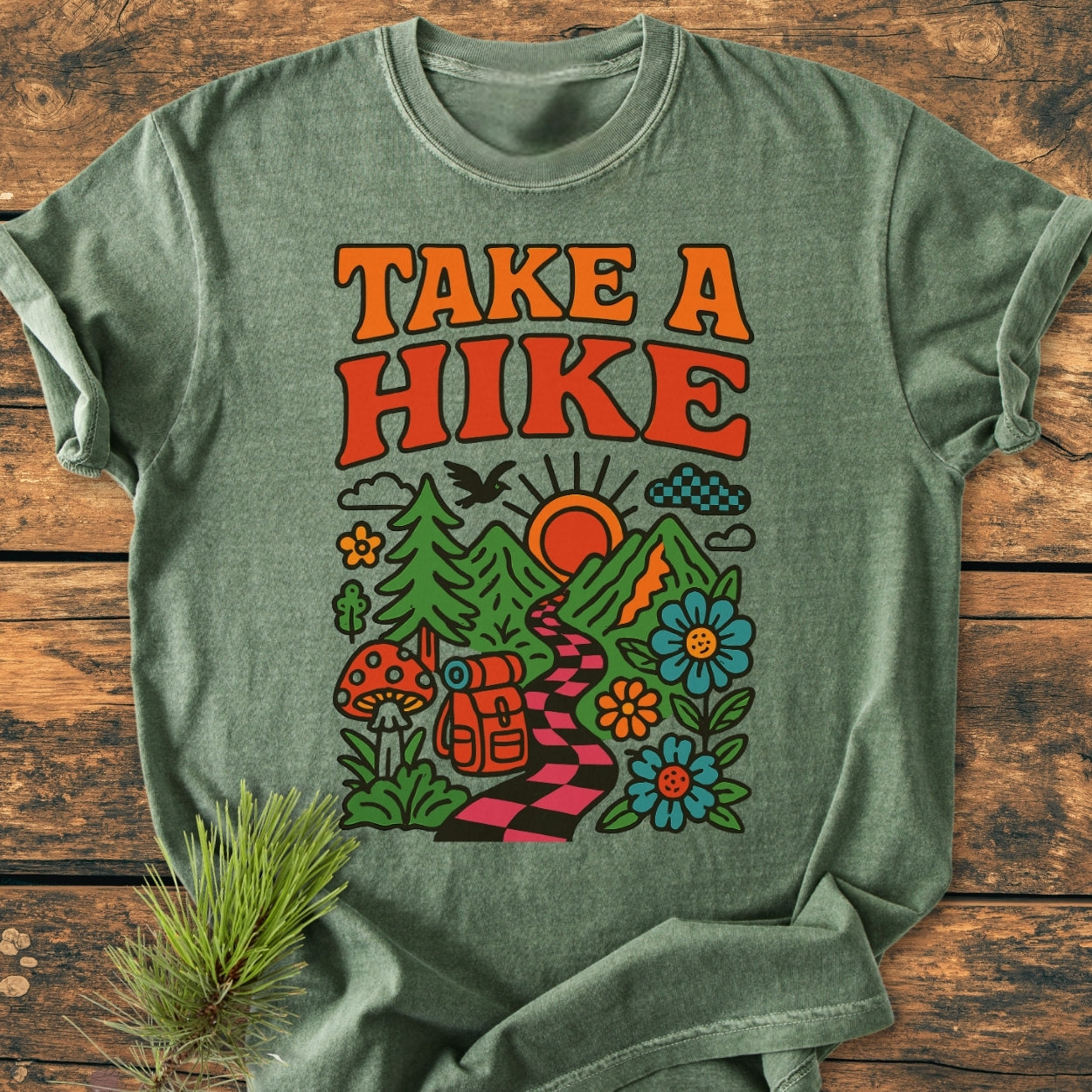 Heather-green Take A Hike Funky - Vintage Wash Tee featuring retro-style text and vibrant nature scene with mountains, trail, and wildlife. Laid on wood with pine sprig, highlighting its relaxed, durable design.