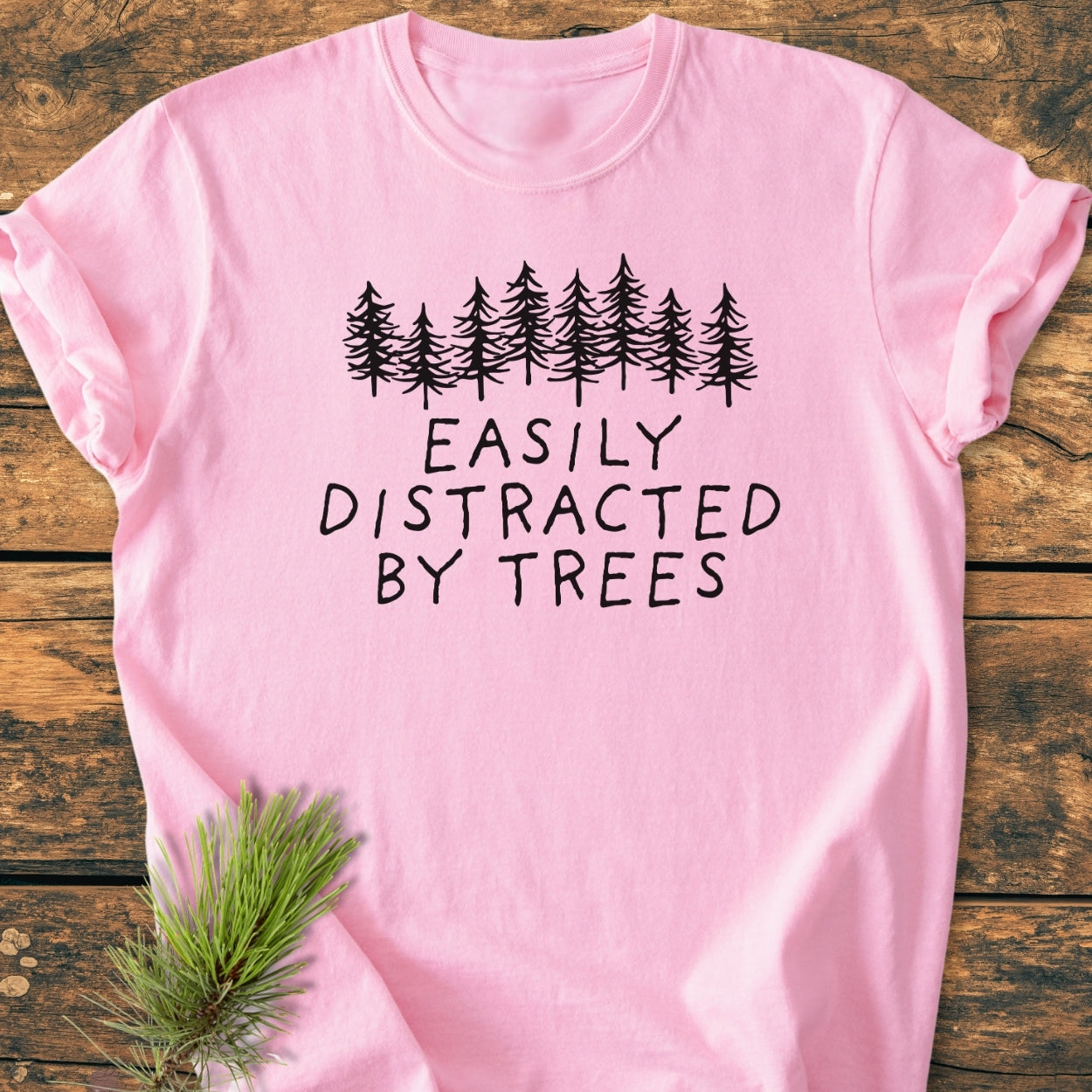 Easily Distracted By Trees - Vintage Wash Tee featuring a black evergreen tree graphic and text on a light-pink garment-dyed shirt, laid flat with pine needles tucked underneath.