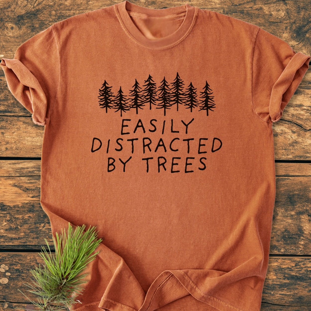 Easily Distracted By Trees - Vintage Wash Tee, featuring pine-tree silhouettes and hand-drawn lettering. Laid flat on a wooden surface with a pine sprig, showcasing its soft, relaxed fit.