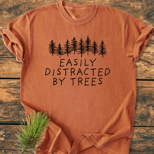 Easily Distracted By Trees - Vintage Wash Tee, featuring pine-tree silhouettes and hand-drawn lettering. Laid flat on a wooden surface with a pine sprig, showcasing its soft, relaxed fit.
