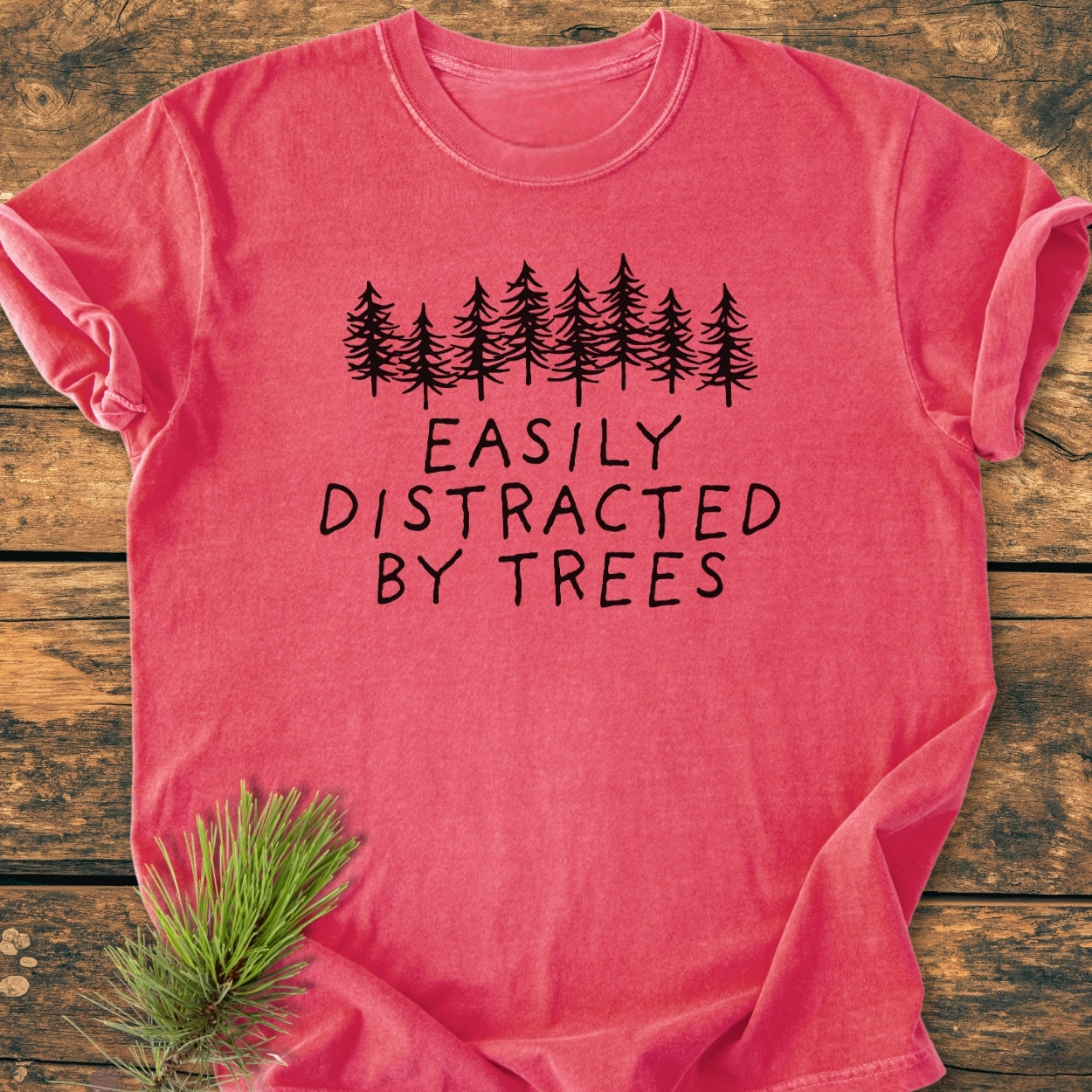 Easily Distracted By Trees - Vintage Wash Tee features black pine-tree silhouettes and slogan on a soft, relaxed-fit t-shirt, laid on wood with pine needles, offering cozy, durable wear.