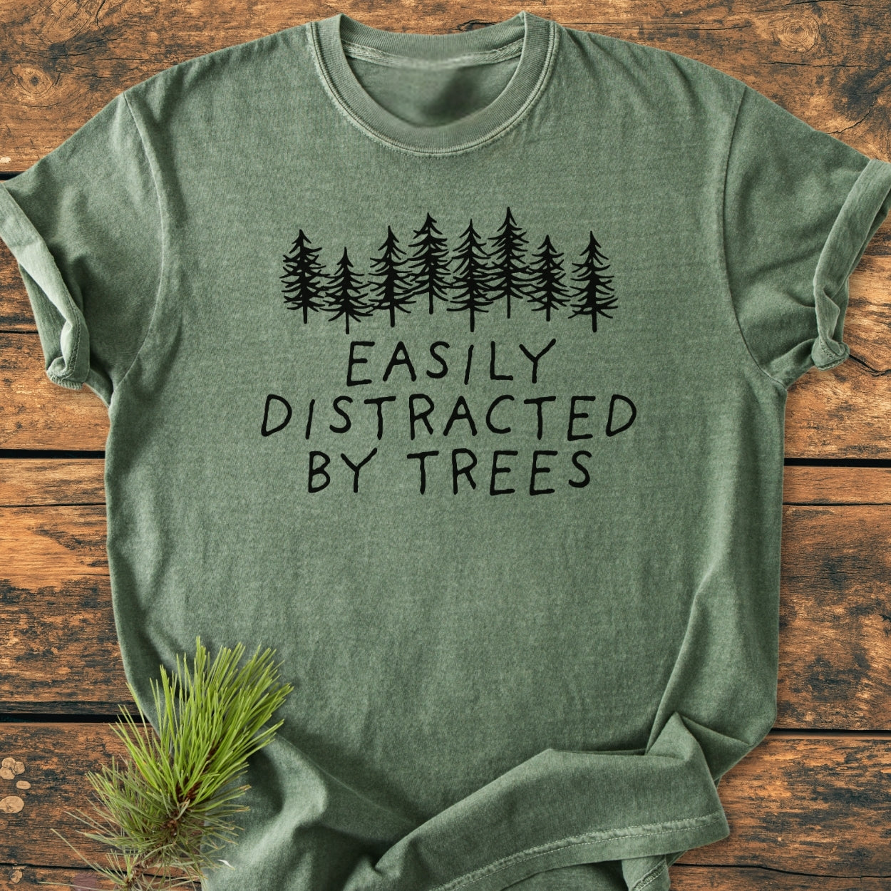 Easily Distracted By Trees - Vintage Wash Tee, featuring a pine tree graphic and slogan, lies on wood. Beside it, a pine branch accentuates the outdoors theme of this heather-green, soft-washed, relaxed-fit shirt.