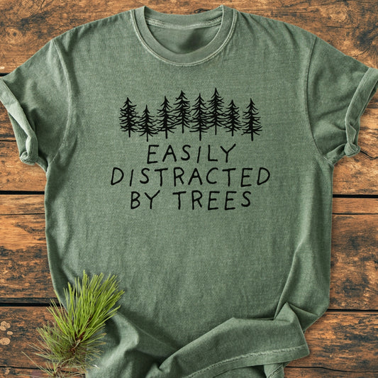 Easily Distracted By Trees - Vintage Wash Tee, featuring a pine tree graphic and slogan, lies on wood. Beside it, a pine branch accentuates the outdoors theme of this heather-green, soft-washed, relaxed-fit shirt.