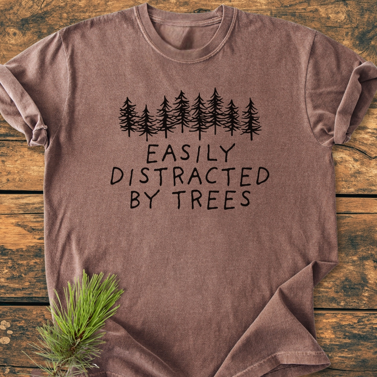 Easily Distracted By Trees - Vintage Wash Tee features stylized black evergreen trees and text on a brown-washed T-shirt, laid flat on a wooden surface with a pine needle sprig nearby.