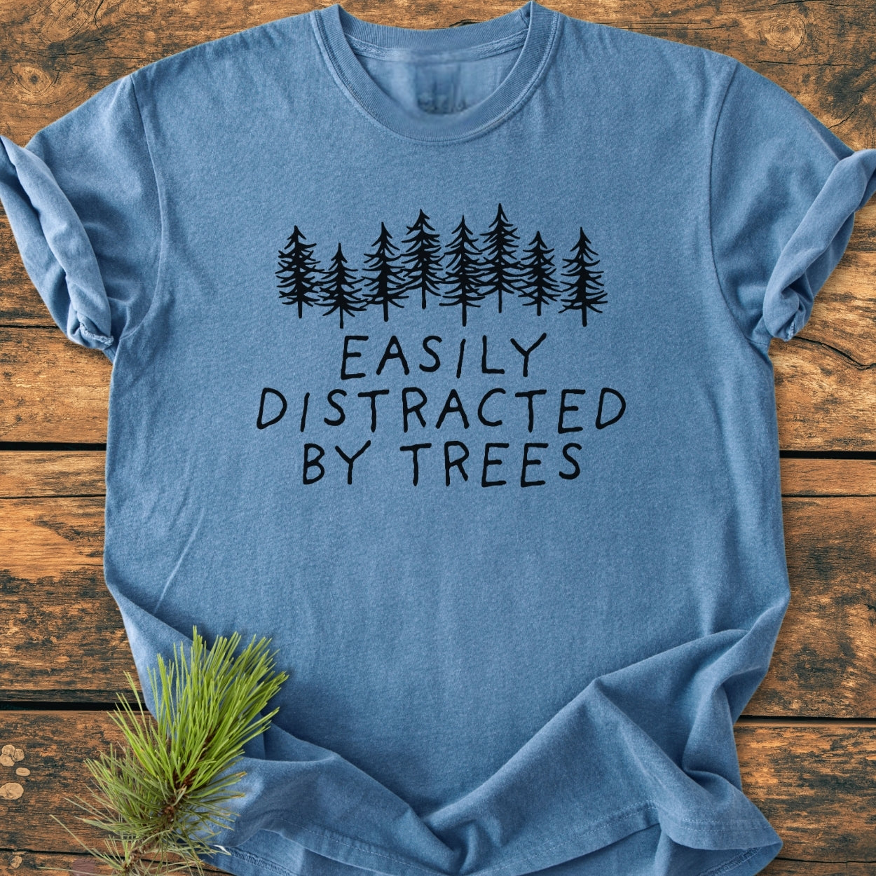 Easily Distracted By Trees - Vintage Wash Tee features a black evergreen tree graphic with text on a heather-blue T-shirt, complemented by a pine branch, highlighting its woodland theme.