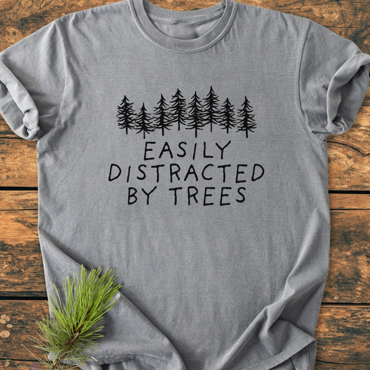 Easily Distracted By Trees - Vintage Wash Tee featuring evergreen tree graphic and slogan, displayed on wooden surface with pine sprig accent. Heather-gray, 100% ring-spun cotton, relaxed fit, durable double-needle stitching.