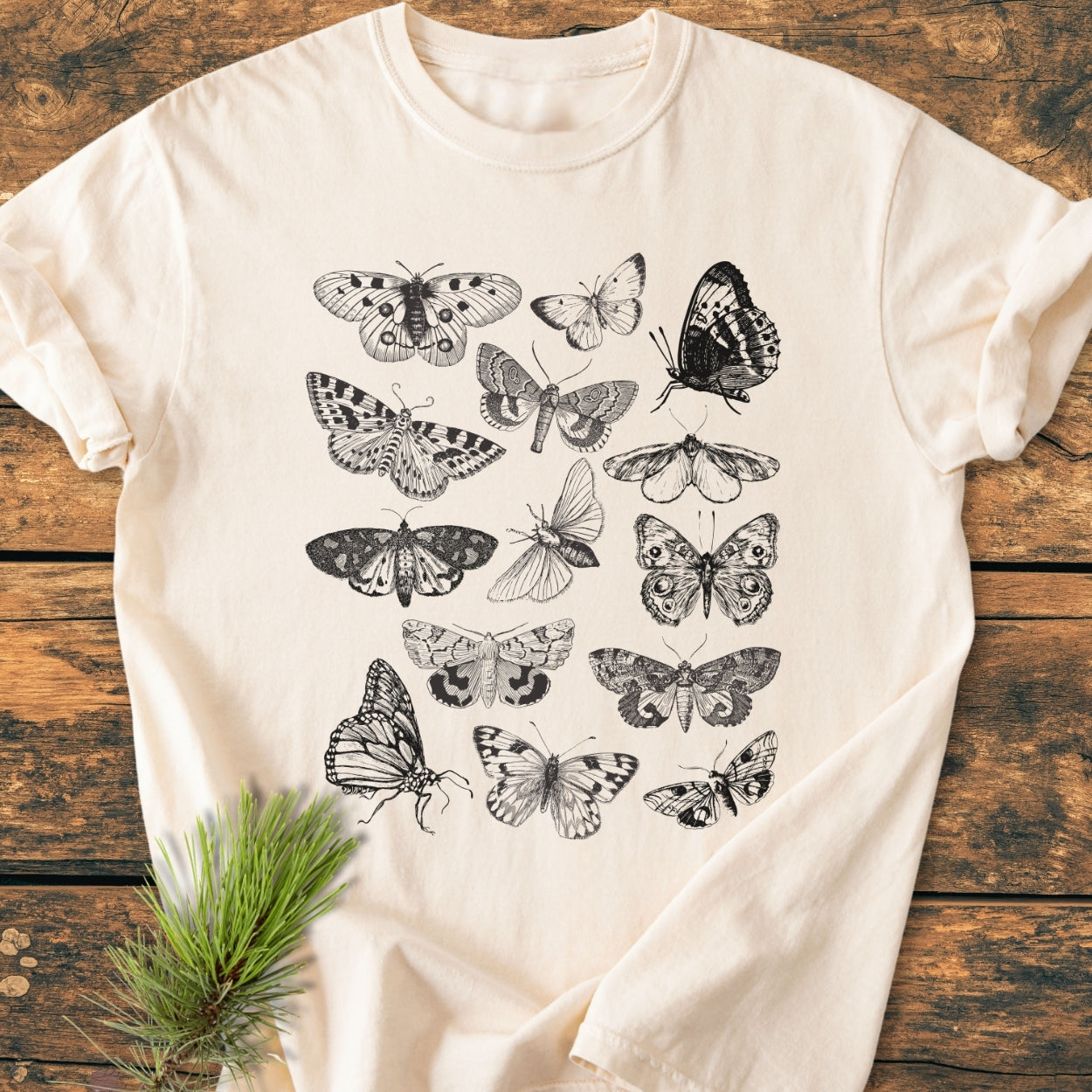 Vintage Butterfly Tattoo - Vintage Wash Tee, light-cream with intricate black-and-white lepidoptera designs, lies on rustic wood. A small pine sprig accents the soft, relaxed fit tee, highlighting its nature-inspired theme.