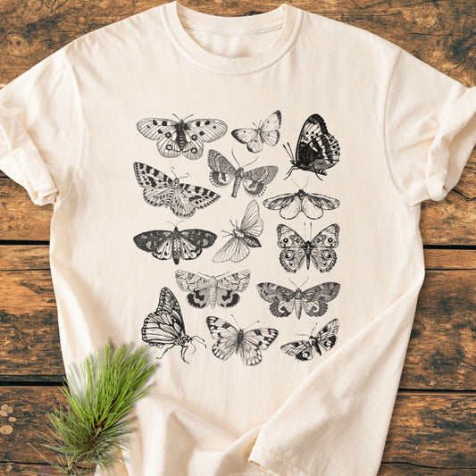 Vintage Butterfly Tattoo - Vintage Wash Tee, light-cream with intricate black-and-white lepidoptera designs, lies on rustic wood. A small pine sprig accents the soft, relaxed fit tee, highlighting its nature-inspired theme.