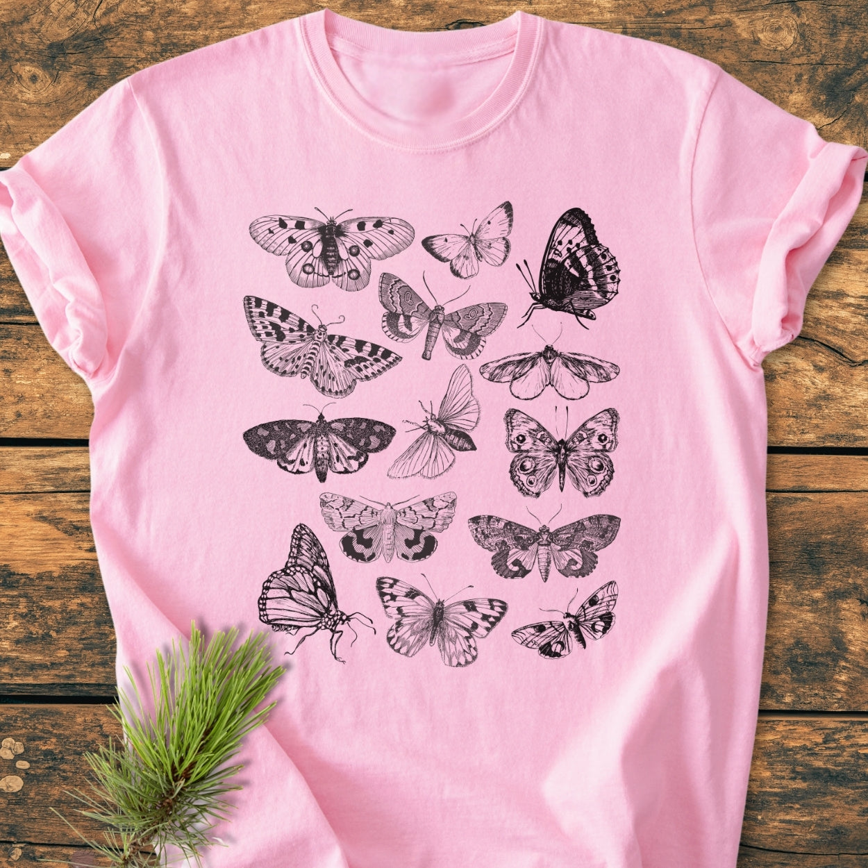 Vintage Butterfly Tattoo - Vintage Wash Tee features hand-drawn moths and butterflies screen-printed in black on a soft pink background, laid flat on wood with a pine sprig accent for a natural touch.