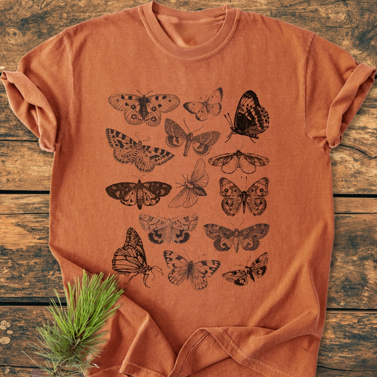 Vintage Butterfly Tattoo - Vintage Wash Tee displayed flat, adorned with black butterfly and moth illustrations in a grid pattern, on a wood surface, complemented by a pine sprig on the hem.