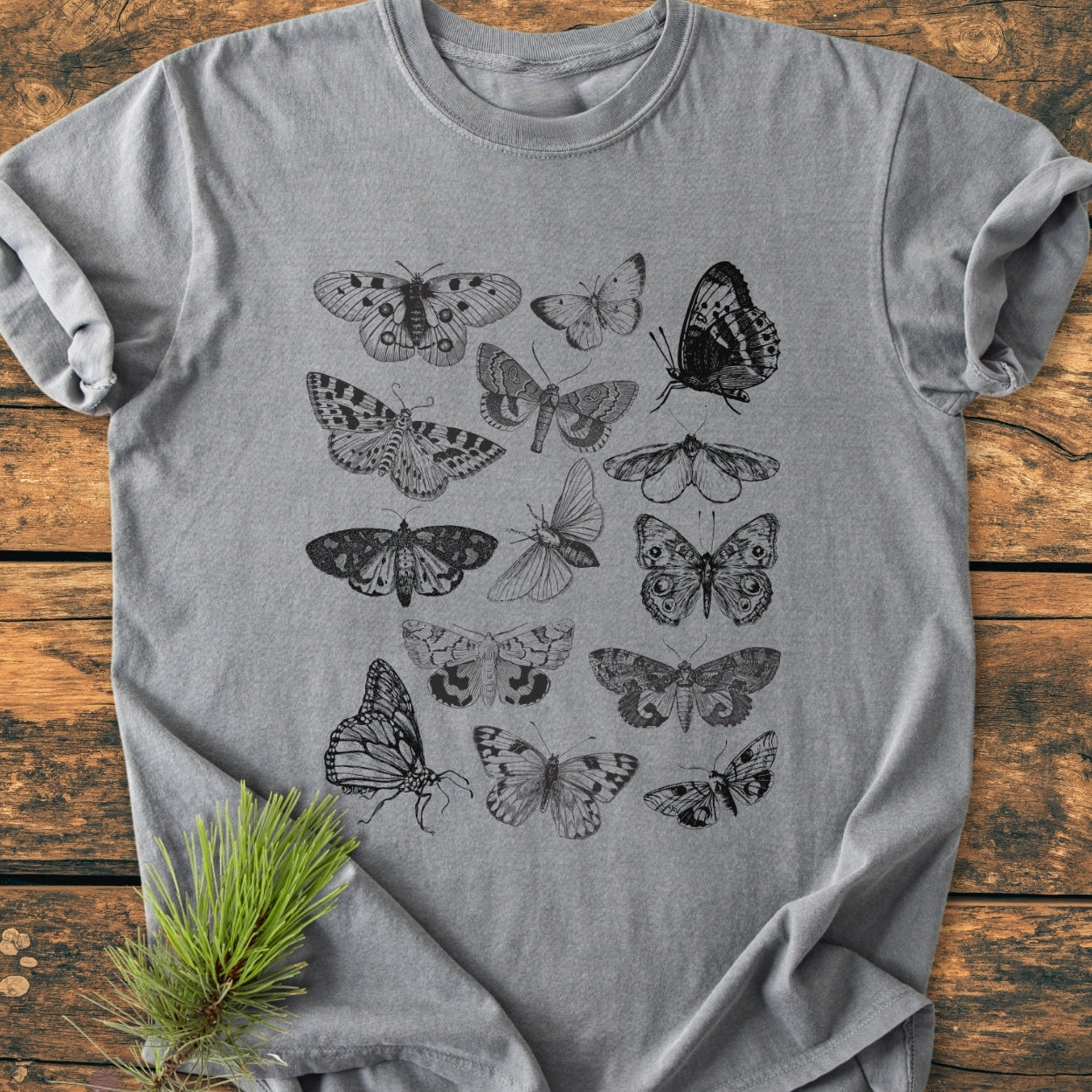 Vintage Butterfly Tattoo - Vintage Wash Tee featuring a grid of black line-drawn butterflies and moths on a heather-gray crew-neck, laid flat with a pine sprig for styling.