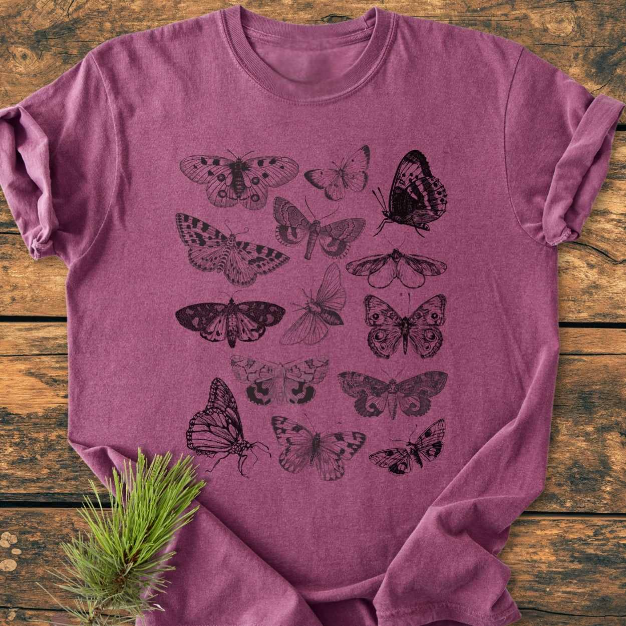 Vintage Butterfly Tattoo - Vintage Wash Tee, featuring hand-drawn butterfly and moth illustrations, displayed folded on a wooden surface with a pine sprig beside it, highlighting the shirt's artistic design.