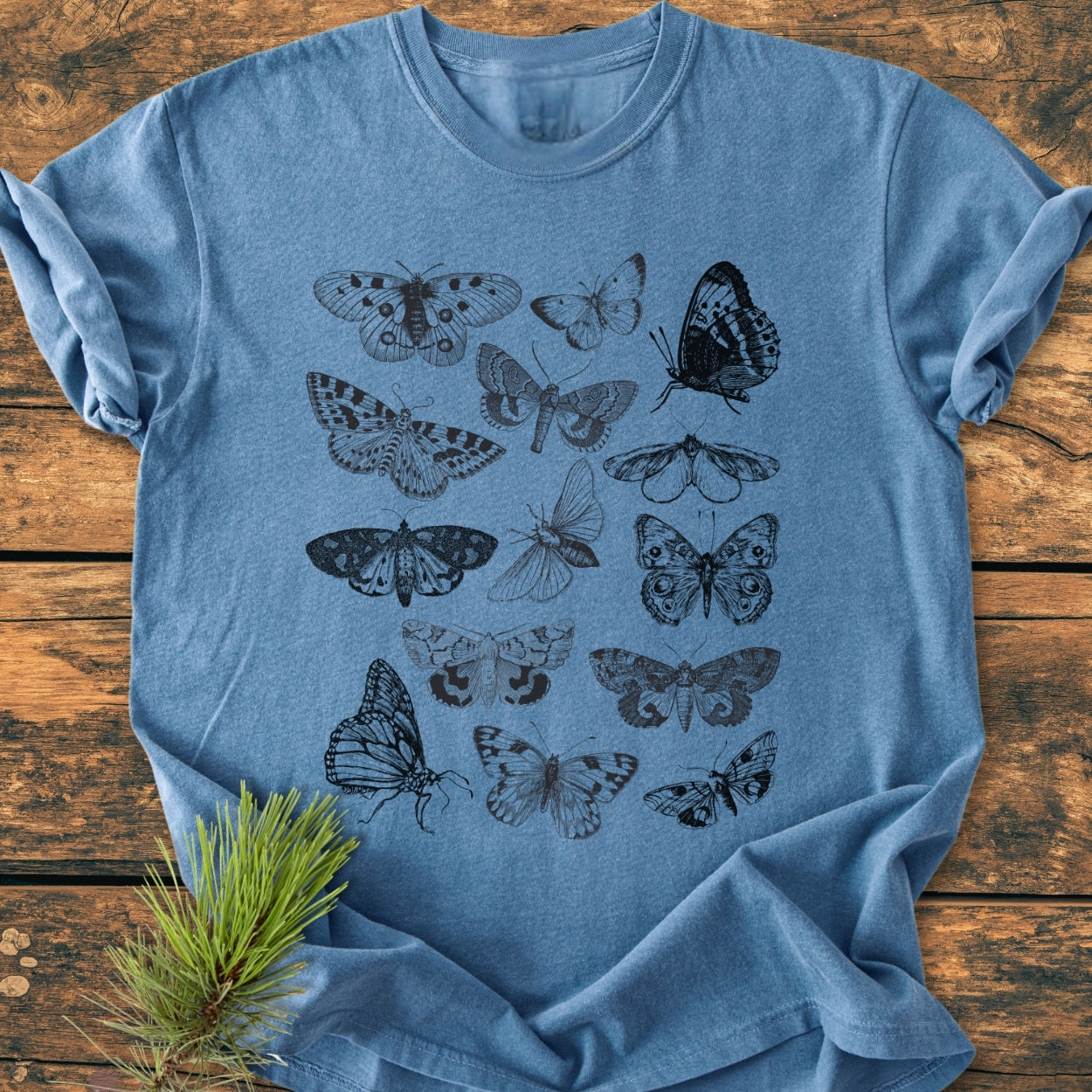 Vintage Butterfly Tattoo - Vintage Wash Tee features intricate black insect illustrations on a heathered blue T-shirt, laid flat with folded sleeves on a wooden surface, emphasizing its detailed design and relaxed fit.