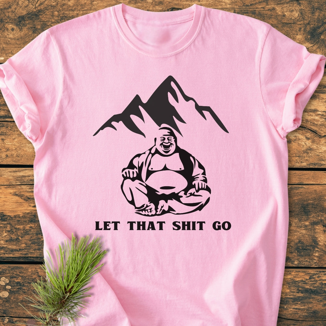 Let That Shit Go - Vintage Wash Tee featuring a laughing Buddha and mountain silhouette, with bold slogan, on a pink T-shirt laid on wood, accompanied by a pine sprig.