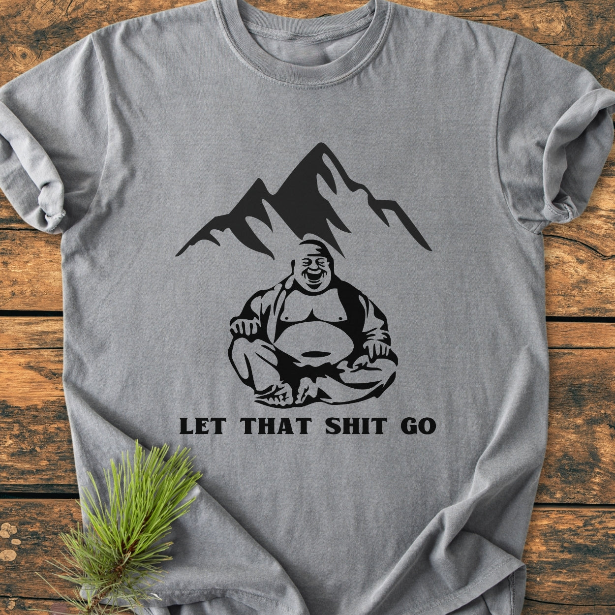 Heather-gray Let That Shit Go - Vintage Wash Tee featuring a laughing Buddha in lotus pose with mountain silhouette, slogan in bold, laid flat on wood with pine sprig beside.