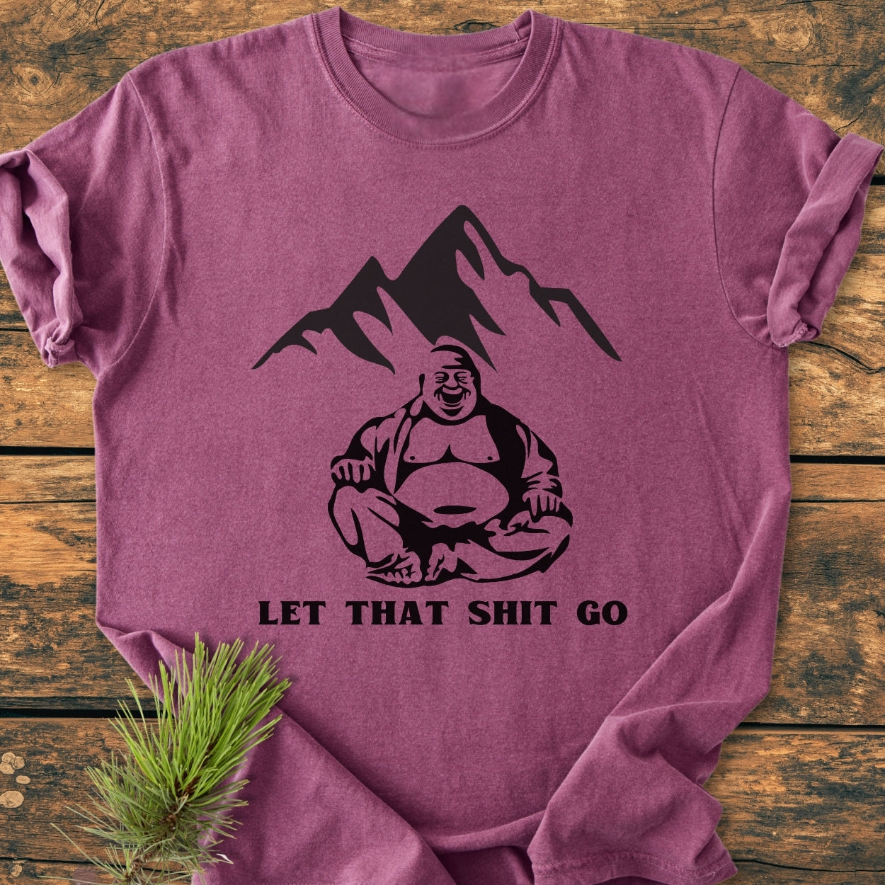 Let That Shit Go - Vintage Wash Tee featuring a stylized smiling Buddha in lotus pose with mountain silhouette, displayed on wood with pine sprig; soft, garment-dyed, relaxed fit for comfort.