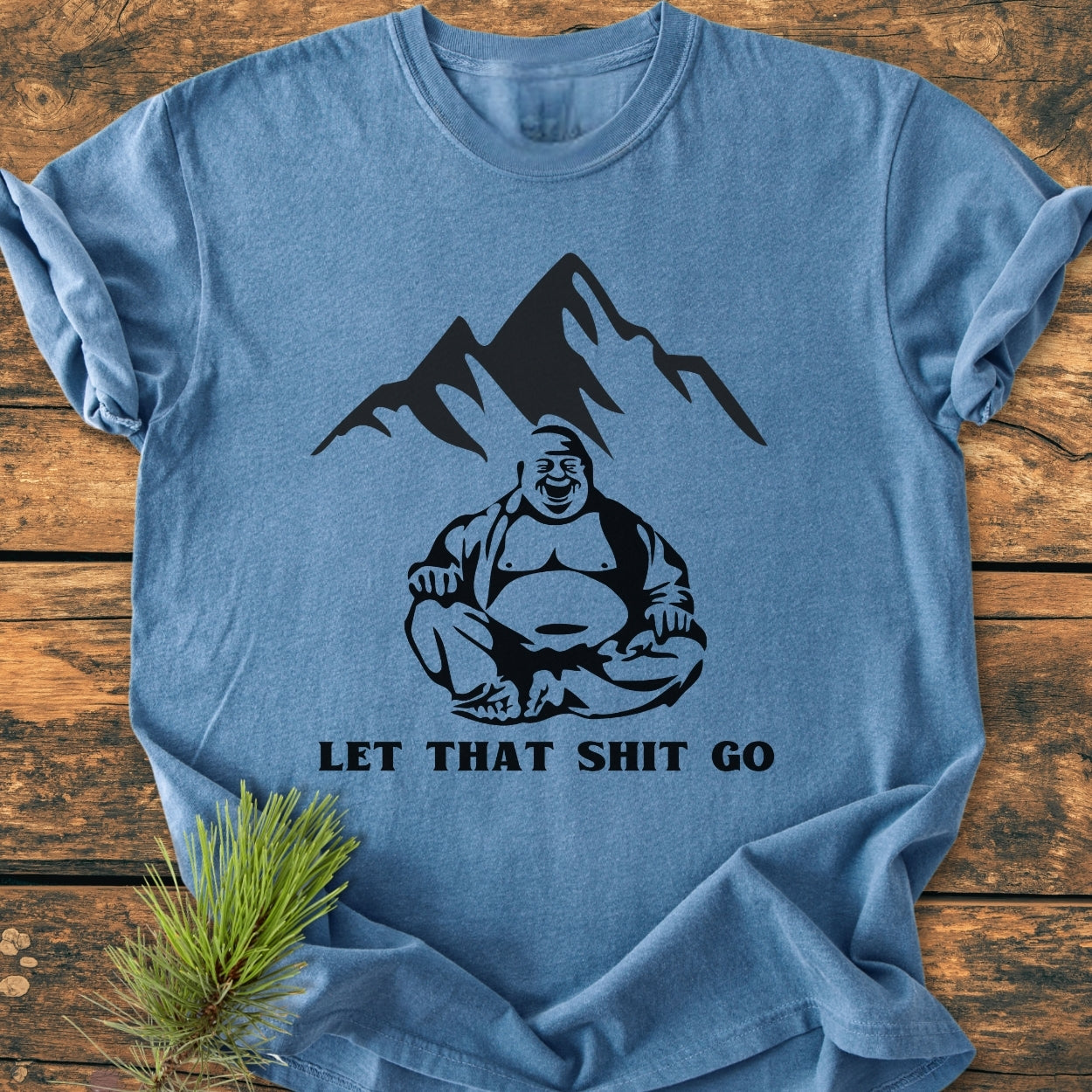 Let That Shit Go - Vintage Wash Tee features a stylized mountain, seated Buddha, and slogan on soft-washed, garment-dyed fabric. Laid on wood, with a pine sprig accent.