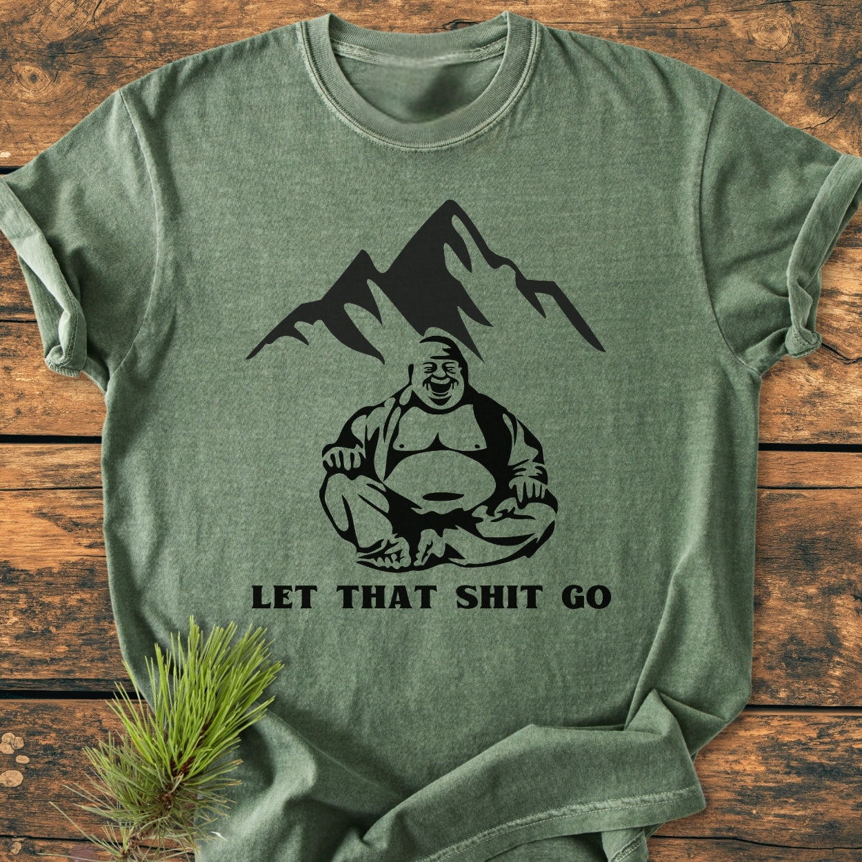 Let That Shit Go - Vintage Wash Tee: Heather-green T-shirt with a seated Buddha graphic and mountains, featuring 'LET THAT SHIT GO' text, laid on rustic wood with pine sprig accentuating its cozy, relaxed fit.