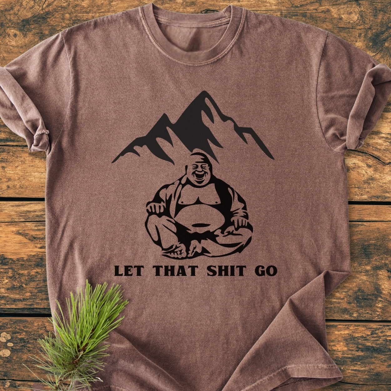 Let That Shit Go - Vintage Wash Tee features a mountain and Buddha graphic with a slogan on a soft, brown, short-sleeve T-shirt, resting on a wooden surface with a pine sprig beside it.