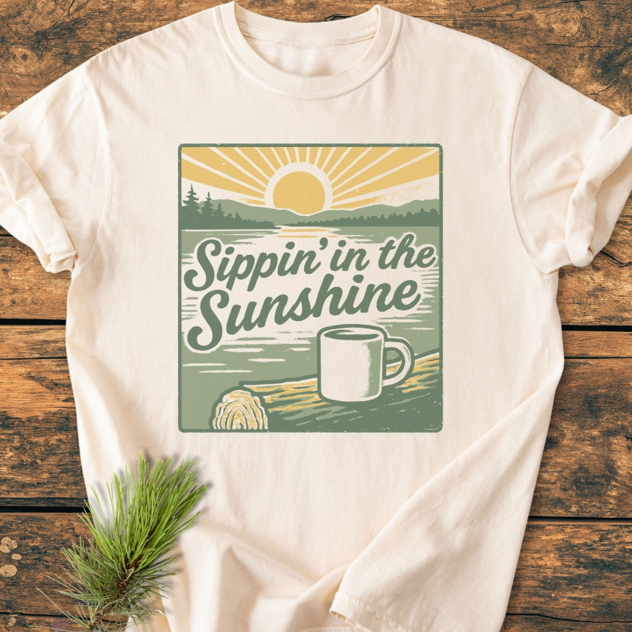 Sippin' In The Sunshine - Vintage Wash Tee with a retro print of a sunrise over a lake, featuring a coffee mug and slogan, displayed on a wooden surface with a pine sprig.