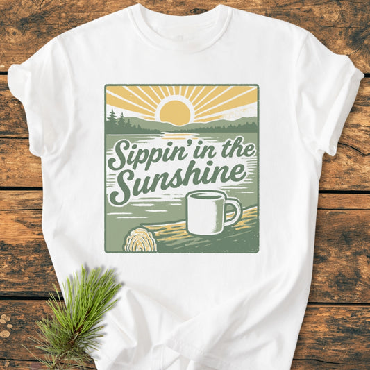 Sippin' In The Sunshine - Vintage Wash Tee, featuring a retro sun and lakeshore scene with a coffee mug. Laid flat on wood, highlighting its soft, durable 100% ring-spun cotton fabric and relaxed fit.