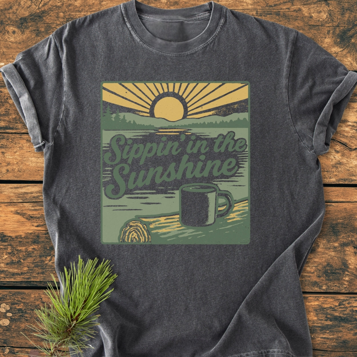 Charcoal-gray Sippin' In The Sunshine - Vintage Wash Tee featuring a retro sunrise graphic over a tree-lined lake, with a cozy coffee scene. Laid on a wooden surface, accented with a pine sprig.