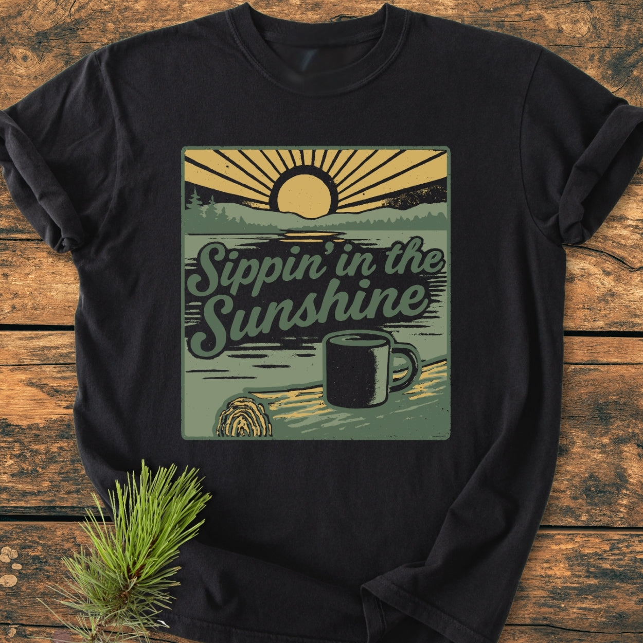 Sippin' In The Sunshine - Vintage Wash Tee featuring a retro sunrise graphic with a coffee mug on driftwood. Displayed flat-lay on wood, accompanied by a pine sprig for a rustic touch.