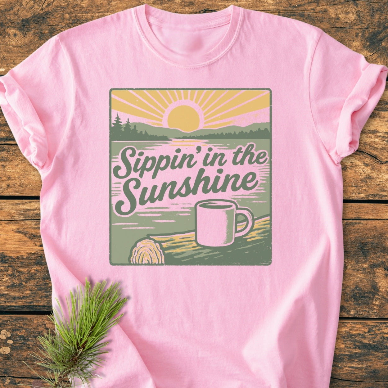 Sippin' In The Sunshine - Vintage Wash Tee features a retro sunrise graphic with a coffee mug and pine trees, on a soft pink T-shirt laid on a wood-grain surface.