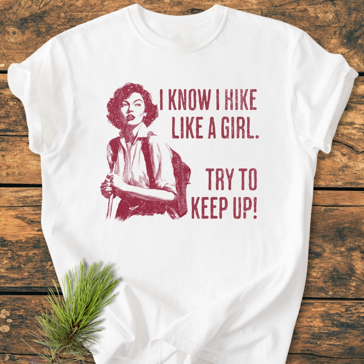 Hike Like A Girl - Vintage Wash Tee featuring a red vintage-style illustration of a woman hiking with bold text. Laid on a wooden surface with a pine sprig accent.