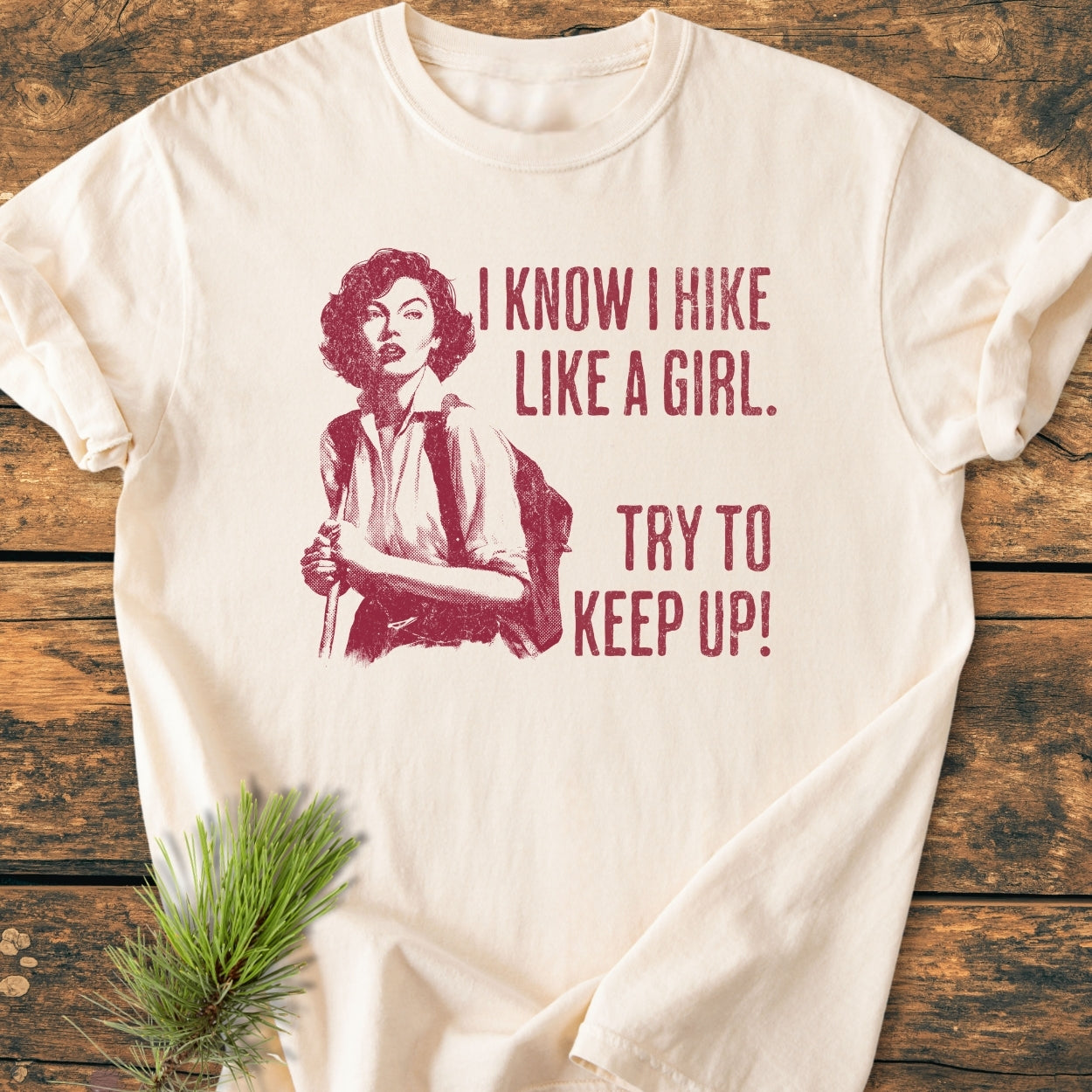 Hike Like A Girl - Vintage Wash Tee featuring a vintage-style illustration of a woman hiking with poles, accompanied by bold motivational text. Laid on wood with an evergreen sprig for a rustic touch.