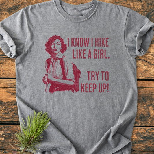 Heather-gray Hike Like A Girl - Vintage Wash Tee on wood, featuring a red graphic of a backpacked woman and slogan. Pine sprig beside. Relaxed fit, 100% ring-spun cotton.