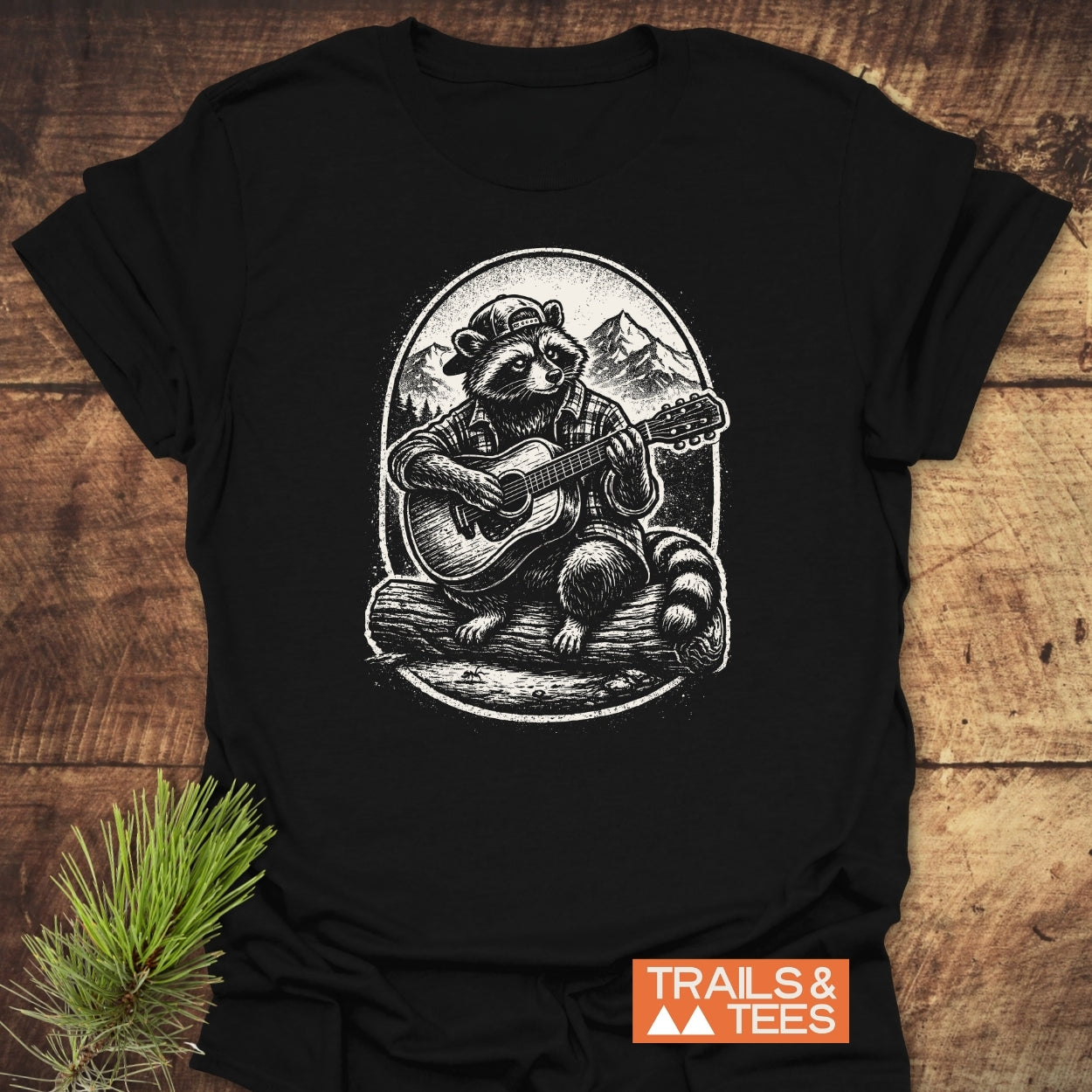 Guitar Playing Raccoon T-Shirt features a raccoon in a cap and plaid shirt, playing guitar on a log, with mountain scenery in the background. Perfect for outdoor enthusiasts seeking comfort and style.