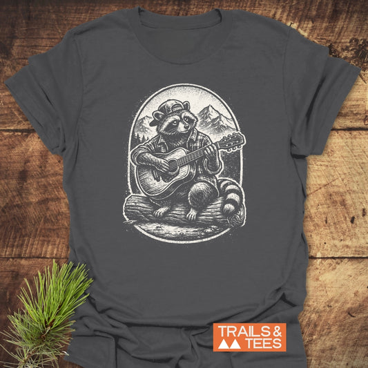 Guitar Playing Raccoon T-Shirt featuring a raccoon in a baseball cap and plaid shirt, playing guitar on a log with mountain backdrop. Displayed with TRAILS & TEES logo and pine needle accent.