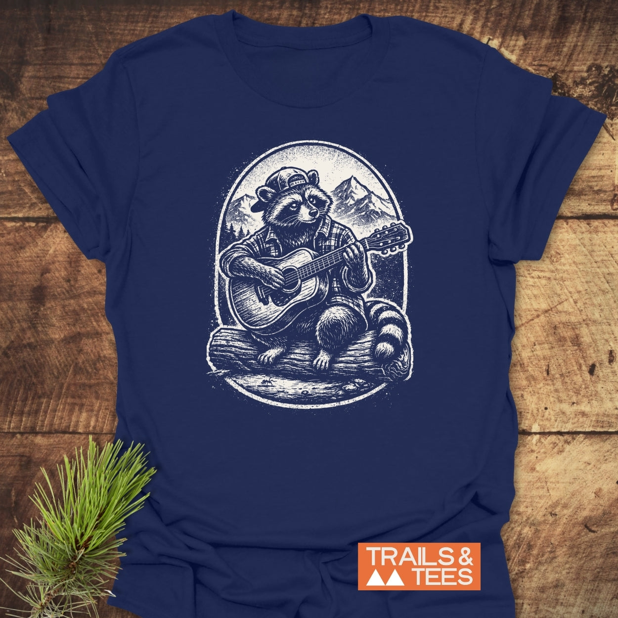 Guitar Playing Raccoon T-Shirt featuring a raccoon in a backward cap and flannel, playing guitar on a log against a mountain backdrop, embodies outdoor adventure. Includes TRAILS & TEES logo.