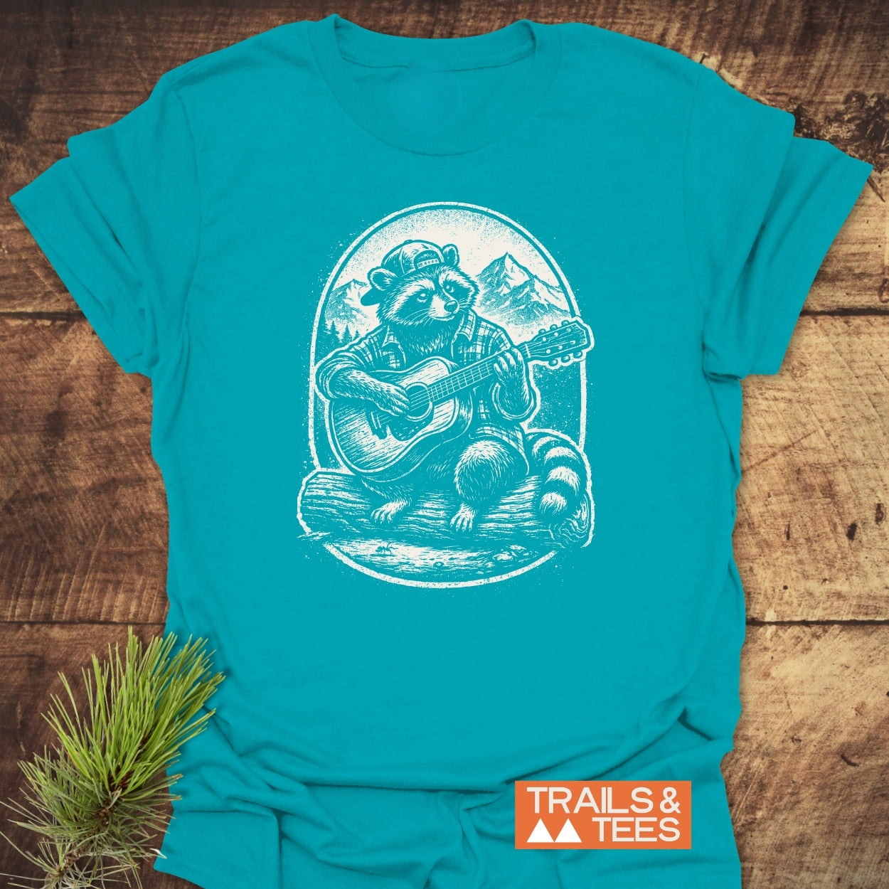 Guitar Playing Raccoon T-Shirt features a raccoon in a cap, playing guitar on a log with mountain backdrop, printed on a teal T-shirt, emphasizing outdoor adventure and comfort. Brand: TRAILS & TEES.