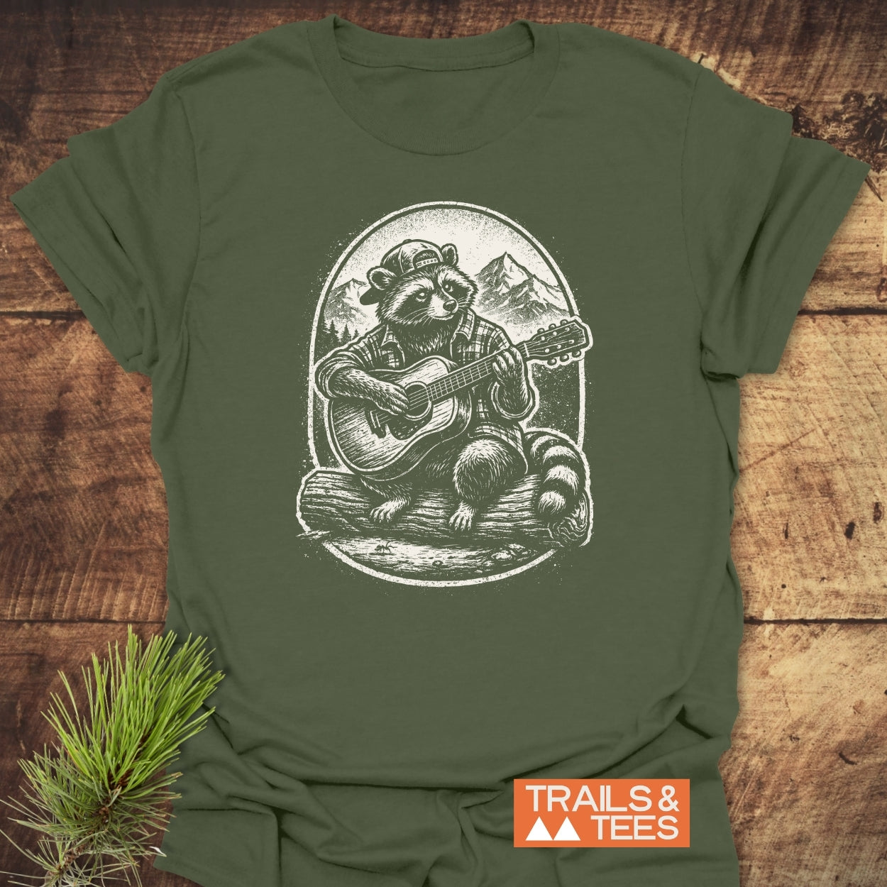 Guitar Playing Raccoon T-Shirt features a raccoon in a backwards cap and flannel, seated on a log, strumming a guitar against a mountain backdrop. Includes TRAILS & TEES logo.
