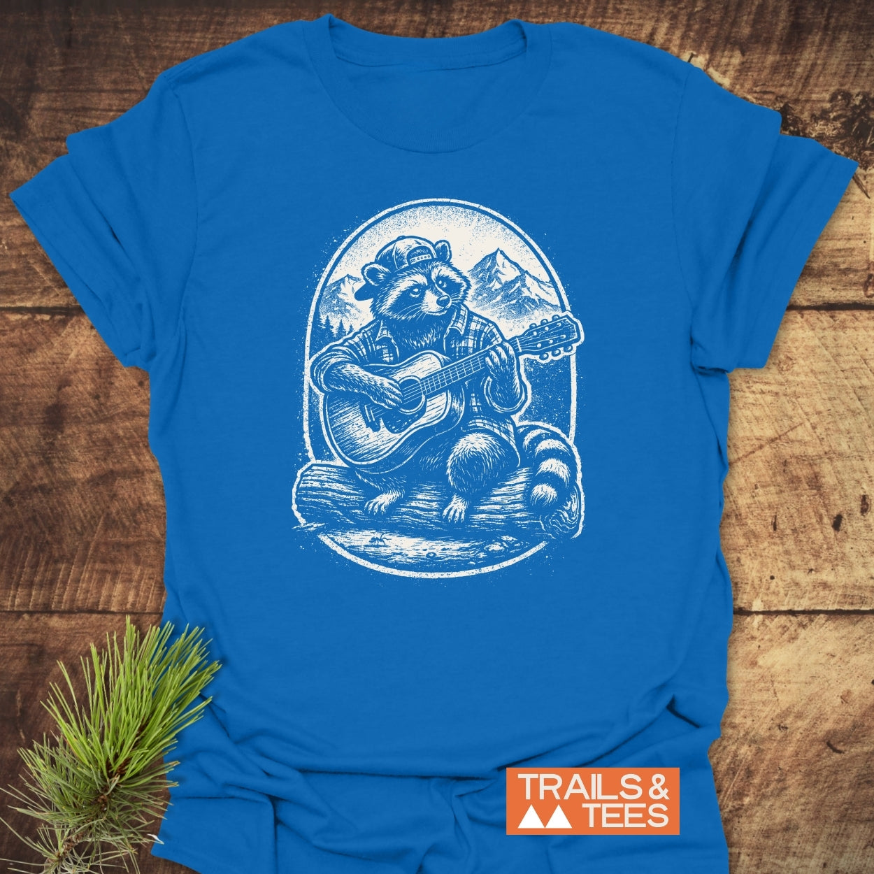 Guitar Playing Raccoon T-Shirt features a raccoon in a plaid shirt and cap, strumming a guitar on a log. Made of soft ring-spun cotton with durable prints, ideal for outdoor lovers.