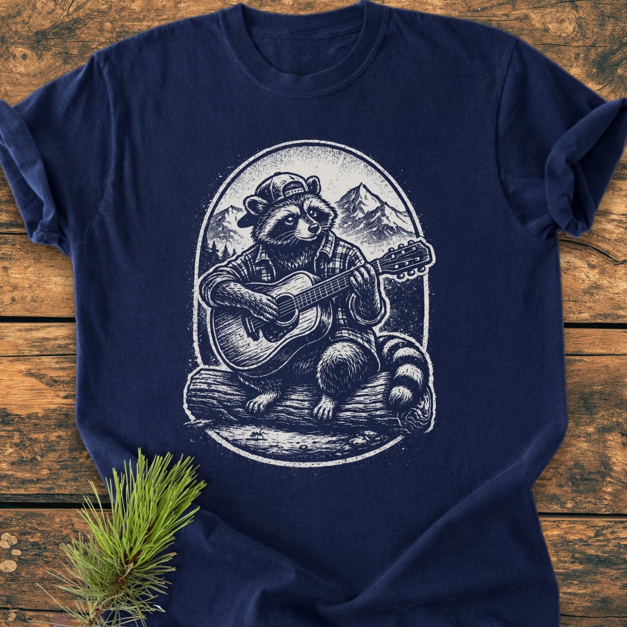 Guitar Playing Raccoon - Vintage Wash Tee features a raccoon in a cap playing guitar, illustrated on a navy-blue T-shirt, laid on rustic wood with a pine branch nearby.