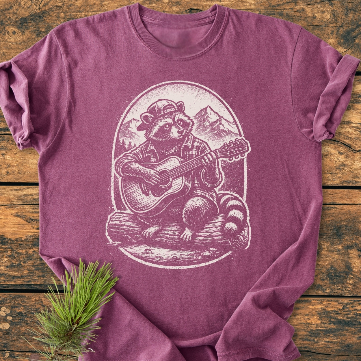 Guitar Playing Raccoon - Vintage Wash Tee features a whimsical raccoon illustration on heather-purple fabric, laid on wooden boards, styled with a pine sprig for a rustic touch.