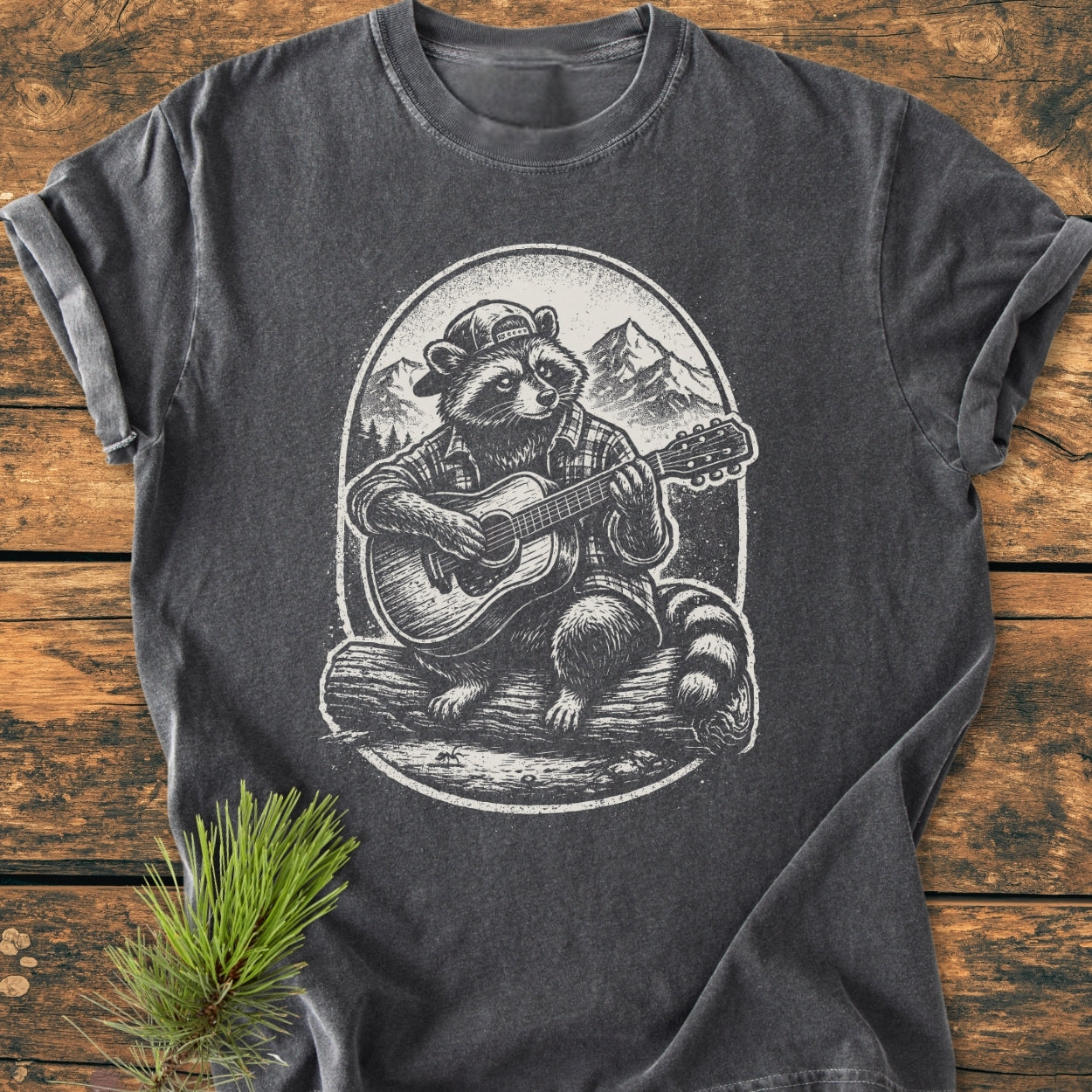 Guitar Playing Raccoon - Vintage Wash Tee: Charcoal-gray T-shirt featuring a raccoon with a cap and plaid shirt playing guitar against mountain and forest backdrop, laid flat on wooden surface with pine sprig.