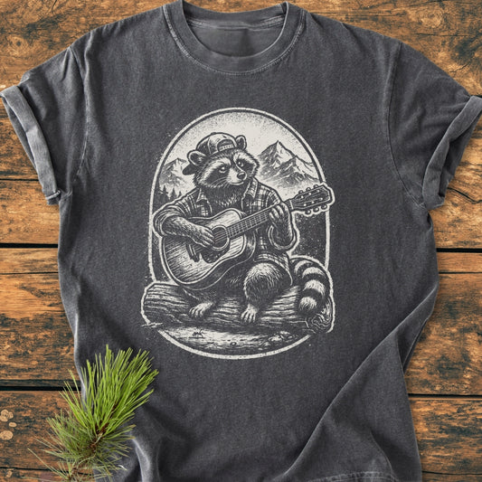 Guitar Playing Raccoon - Vintage Wash Tee: Charcoal-gray T-shirt featuring a raccoon with a cap and plaid shirt playing guitar against mountain and forest backdrop, laid flat on wooden surface with pine sprig.