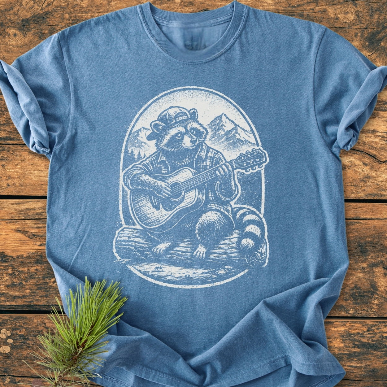 Guitar Playing Raccoon - Vintage Wash Tee featuring a raccoon in a cap and flannel, playing guitar against mountains, laid on wood with a pine sprig beside. Relaxed fit, durable, soft-washed fabric.