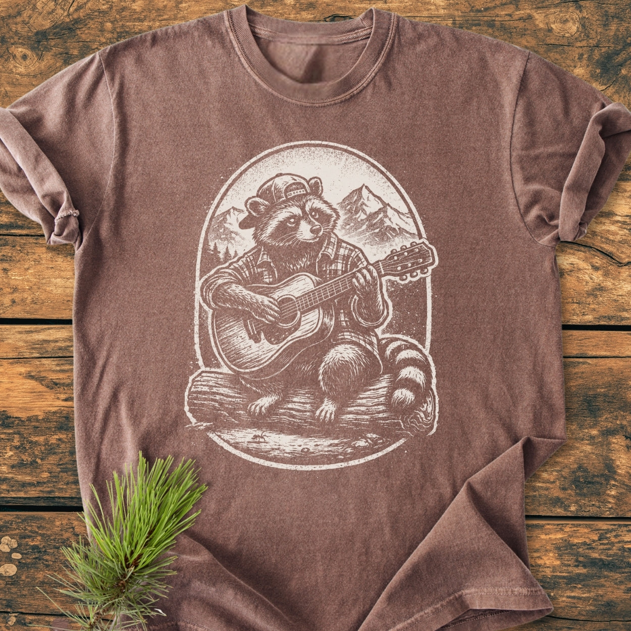 Guitar Playing Raccoon - Vintage Wash Tee features a raccoon in a plaid shirt and cap, strumming a guitar, printed on a faded-brown, short-sleeved T-shirt laid on a rustic wooden surface.