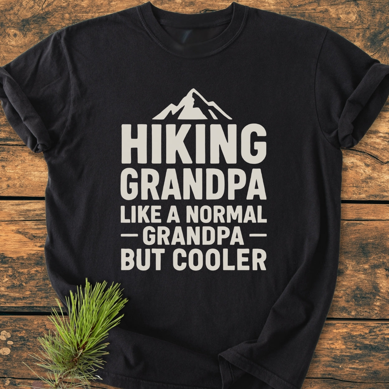 Hiking Grandpa - Vintage Wash Tee displayed on wood with a pine sprig, featuring a white mountain graphic and slogan. Soft-washed, relaxed fit, double-stitched for durability, made of pre-shrunk, ring-spun cotton.
