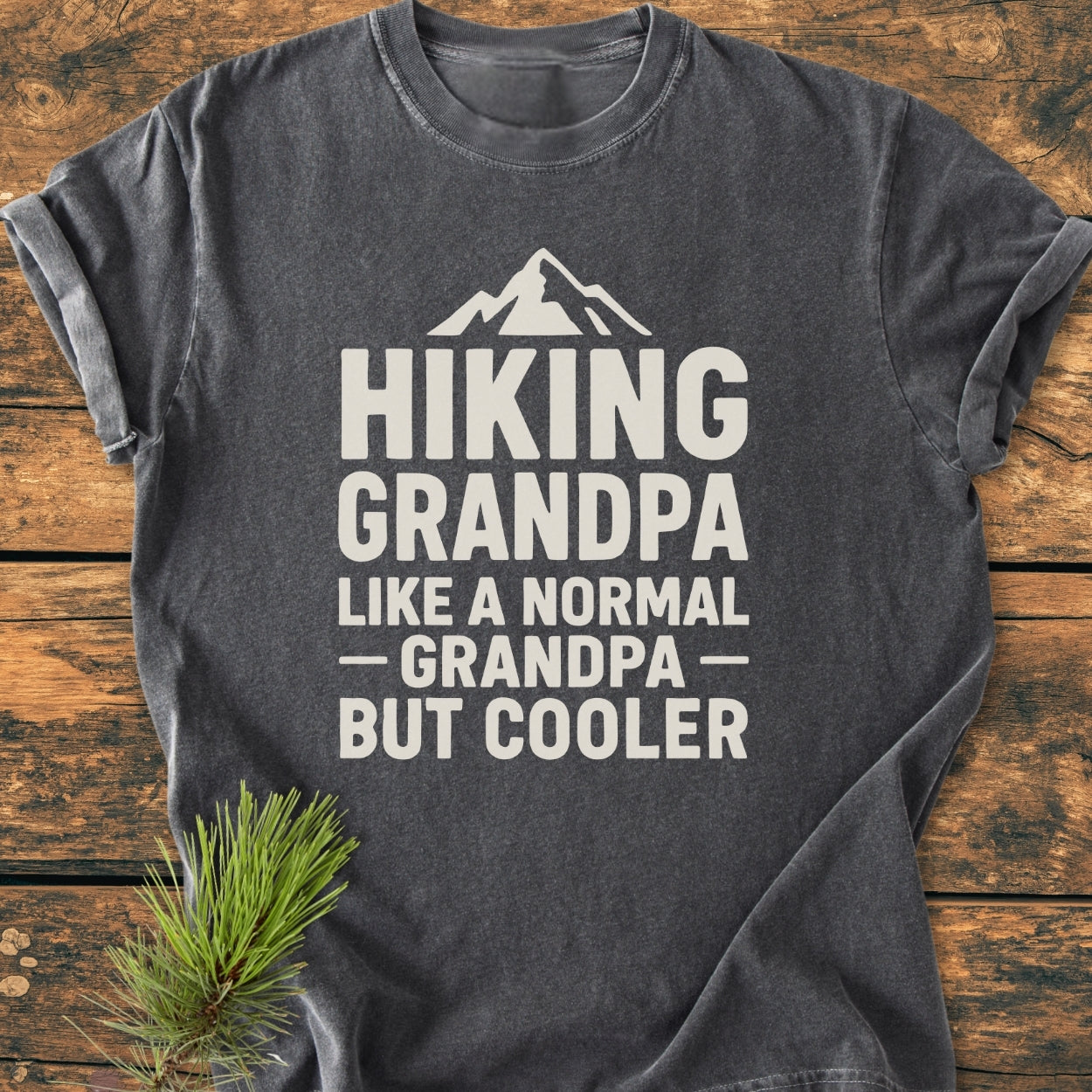 Hiking Grandpa - Vintage Wash Tee features a stylized mountain peak graphic with a catchy slogan on a dark-gray, short-sleeve shirt, laid on wood with a pine sprig nearby.