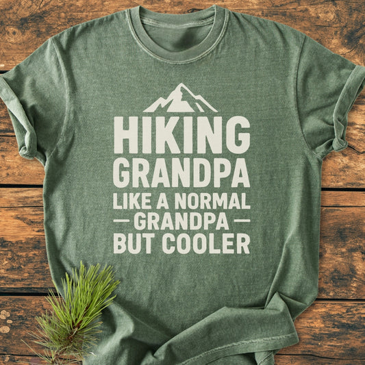 Heather-green Hiking Grandpa - Vintage Wash Tee on rustic wood, featuring a mountain graphic and bold text. Pine needles accentuate the hiking theme. Soft, relaxed fit highlights comfort and durability.