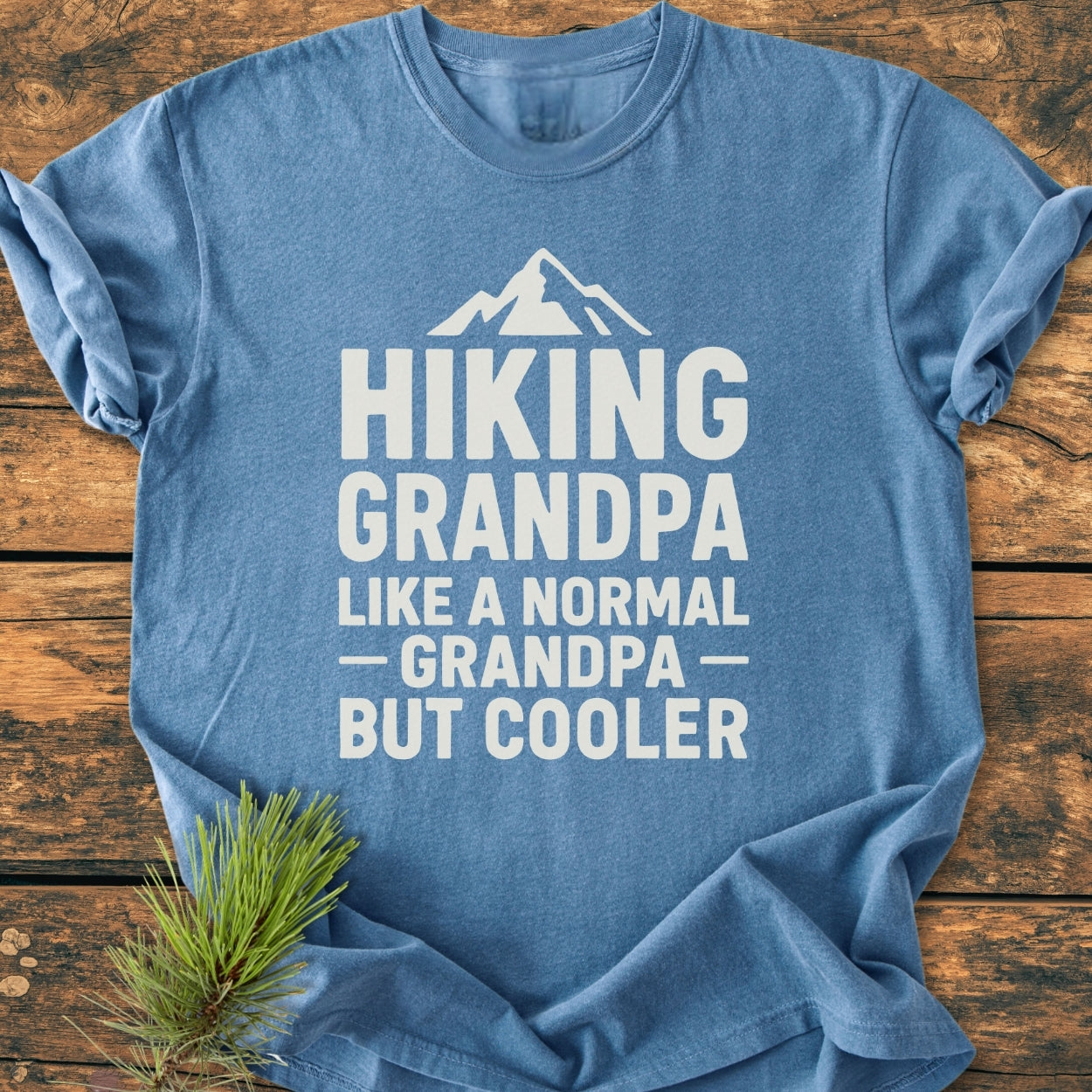 Heather-blue Hiking Grandpa - Vintage Wash Tee with white text and mountain silhouette, laid on rustic wood, accompanied by a small pine sprig. Cozy, relaxed fit, 100% ring-spun cotton.