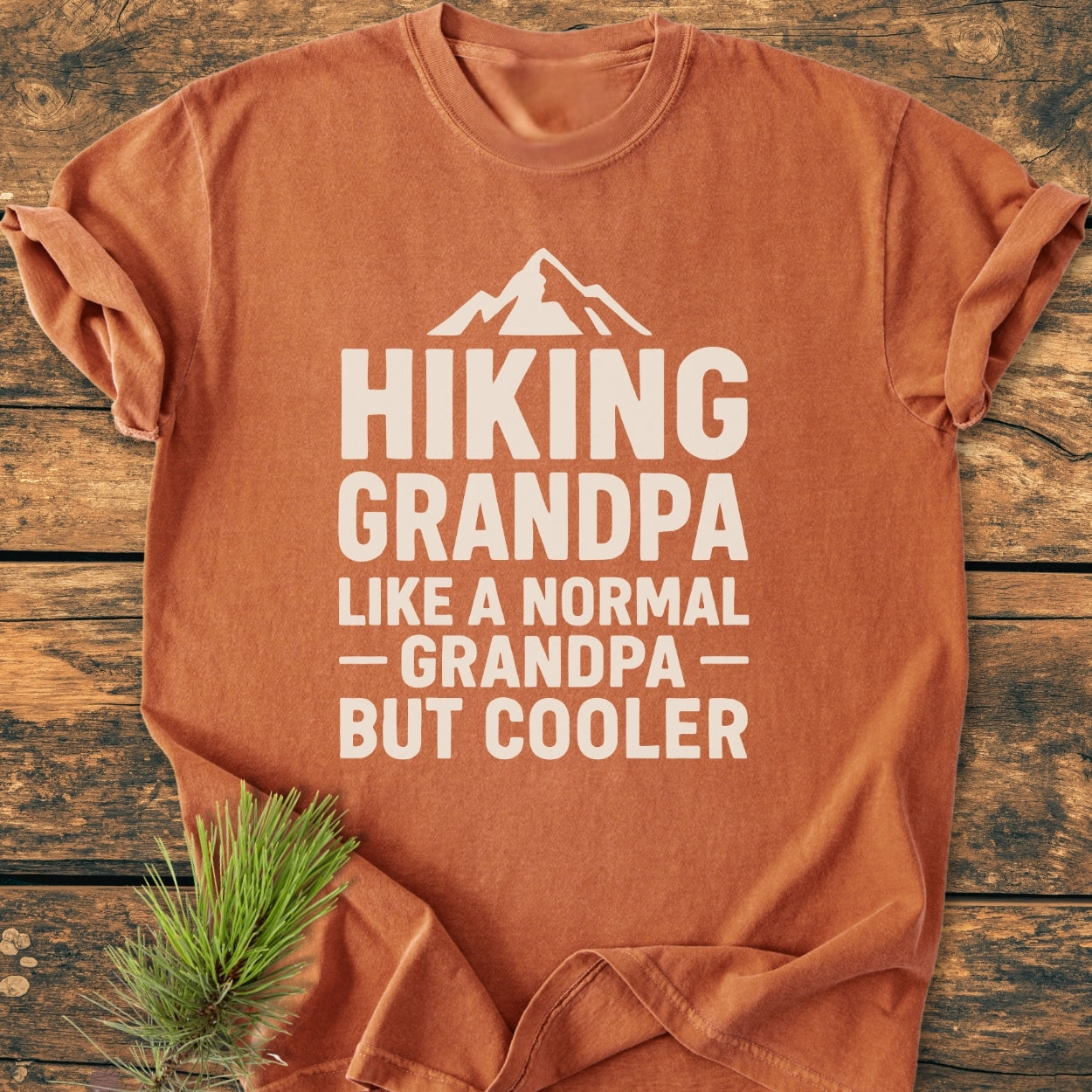 Folded burnt-orange Hiking Grandpa - Vintage Wash Tee with a white mountain graphic and text, styled on wood with pine. Made of soft garment-dyed cotton for comfort.