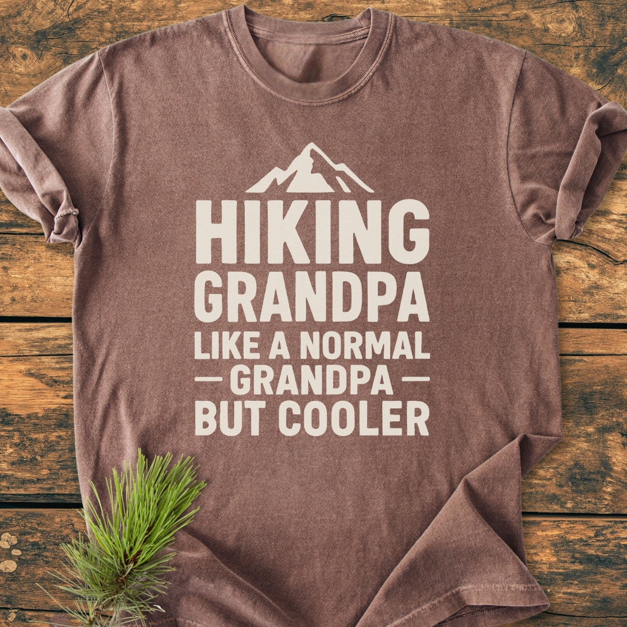 Hiking Grandpa - Vintage Wash Tee in brown with a white mountain graphic and bold text, displayed on wood. Features include short sleeves, 100% cotton, relaxed fit, and decorative pine sprig accent.